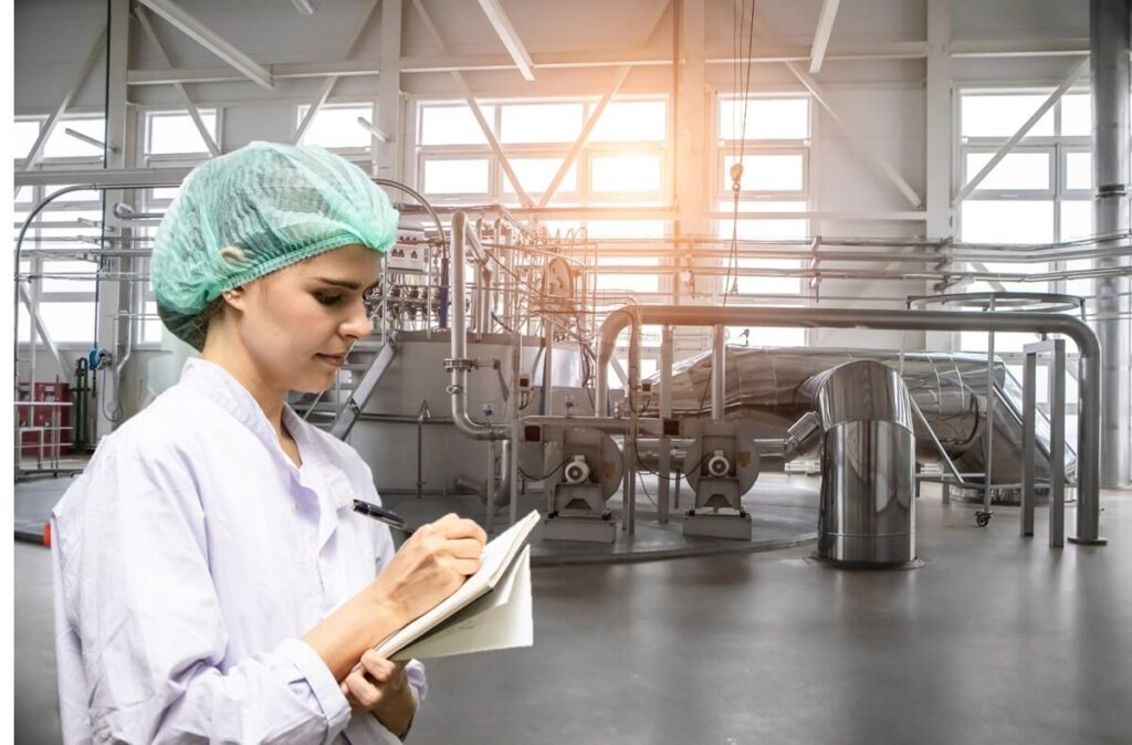 Quality control personnel taking notes inside a powdered milk processing facility, with stainless steel equipment in the background.