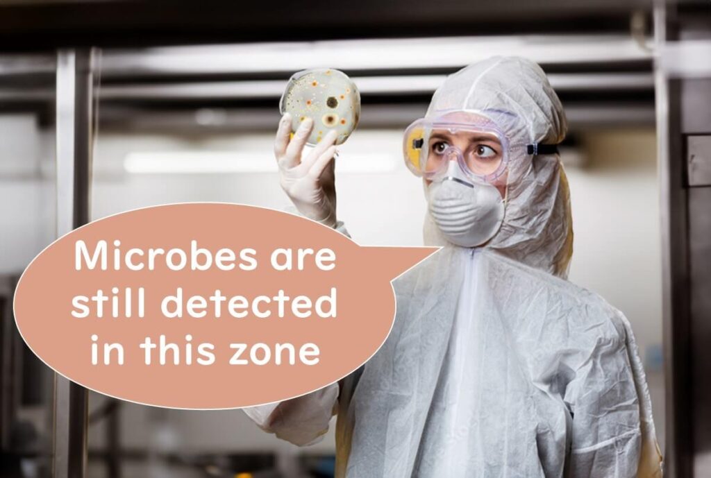 Scientist in full protective clothing examining a petri dish, with a speech bubble stating “Microbes are still detected in this zone,” highlighting microbial presence even in high hygiene areas.